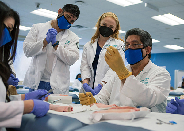 A group of NSU students wearing white coats, masks, and gloves looking over a professor's shoulder. He is holding an unidentified specimen. 
